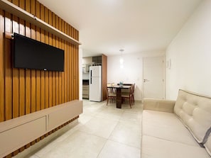A living room with a flat-screen TV mounted on a wooden panel wall, a dining area with a table and chairs, and a sofa. - Puerto By Hosppedar (João Pessoa)