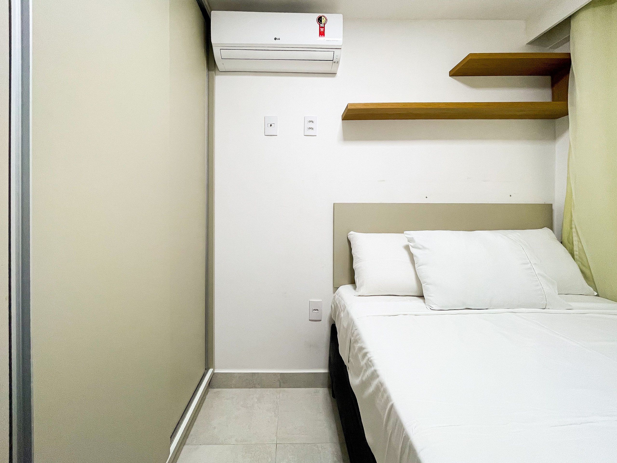 A small, single-bed room with a white bed, a wooden shelf above the bed, and a wall-mounted air conditioner.