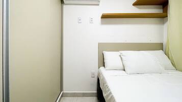 A small, single-bed room with a white bed, a wooden shelf above the bed, and a wall-mounted air conditioner.