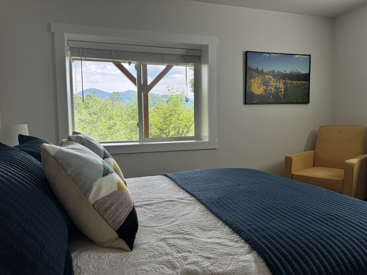A bedroom with a bed, pillows, a chair, a window with a view, and a framed picture on the wall.