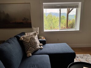 A living room with a blue sofa, a patterned throw pillow, a window with a view of greenery, and a painting on the wall. - Mountain Peace 25 Min from Downtown Asheville (Barnardsville)