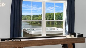 Una habitación con un ventanal grande que da a un lago y un bosque, una mesa de madera con un juego de mesa, y un letrero de neón en la pared.