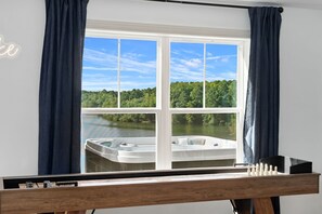 A room with a large window overlooking a lake and forest, a wooden table with a board game, and a neon sign on the wall.