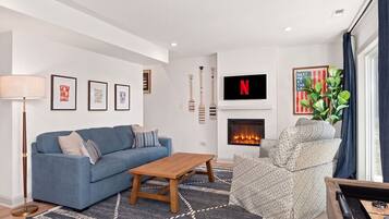 A modern living room with a blue sofa, a wooden coffee table, a patterned armchair, a fireplace, and a mounted TV.