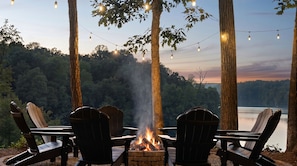 A fire pit with chairs around it, overlooking a lake with trees in the background.