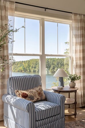 Una habitación con un sillón a rayas, una pequeña mesa redonda con una lámpara, y vistas a un lago y un bosque a través de amplios ventanales.
