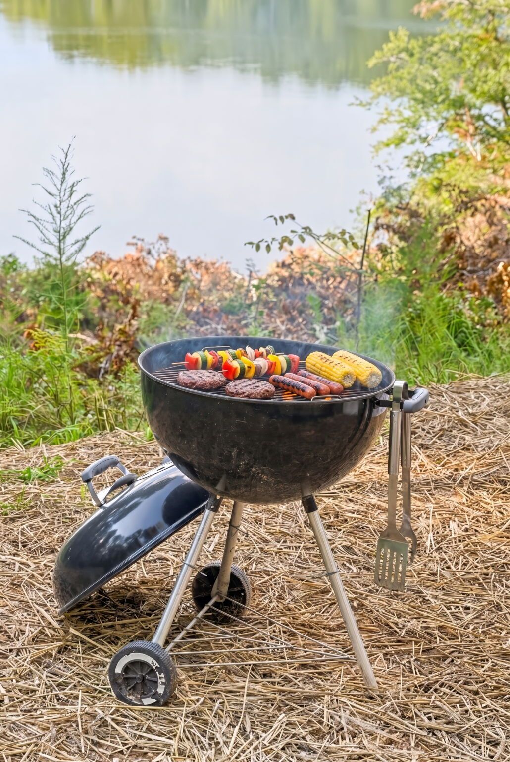 A portable grill with skewers of vegetables and sausages, set on a bed of straw, with a body of water and greenery in the background.