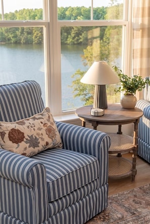 A room with a view of a lake, a striped armchair, a round table with a lamp, and a potted plant.