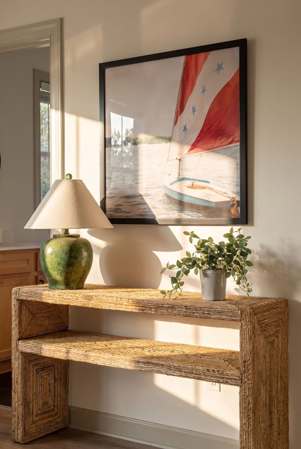 Una consola de madera con una lámpara y una planta en maceta, un cuadro con una imagen de un velero y un espejo en la pared.