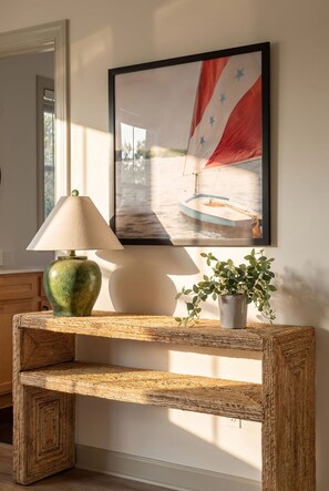 Una consola de madera con una lámpara y una planta en maceta, un cuadro con una imagen de un velero y un espejo en la pared.