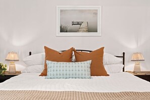 A neatly made bed with white bedding and two brown pillows, flanked by two bedside tables with lamps, against a plain wall with a framed picture above.
