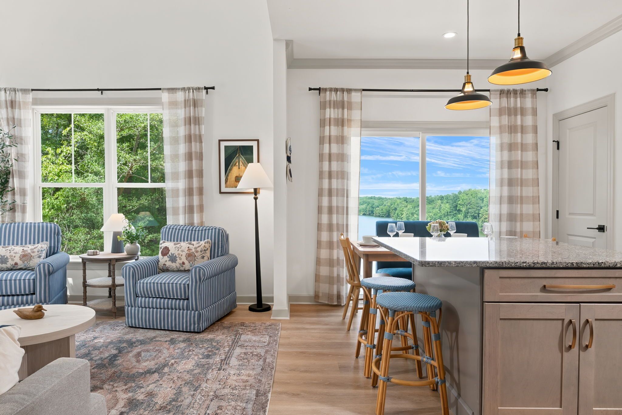 A modern living room with a sofa, armchair, and a dining area with bar stools. There is a large window with a view of greenery.