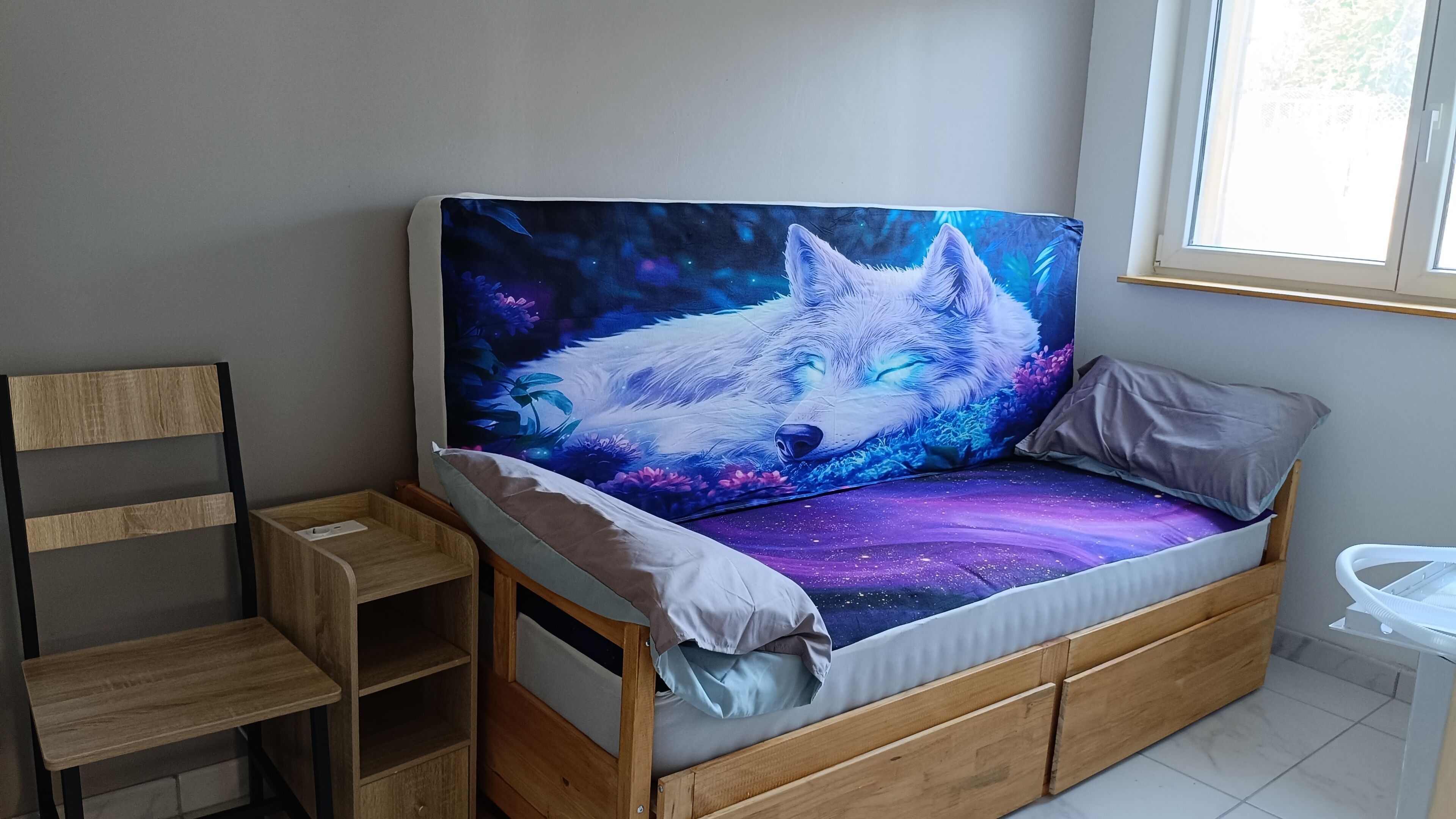Een bed met een matrasovertrek met een ruimte thema, een houten hoofdbord, een klein houten plankje en een raam met daglicht.