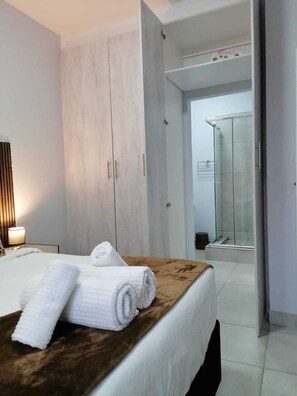 A hotel room with a bed, a wooden headboard, a nightstand with a lamp, and a bathroom visible through an open door.