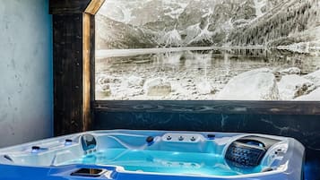 A room with a large window displaying a mountainous landscape, a blue jacuzzi, and a wooden ceiling.