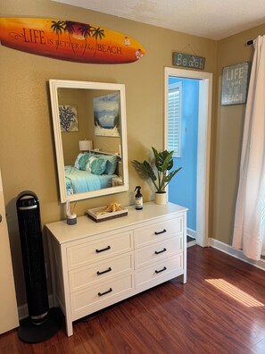Une chambre avec un lit, une commode, un miroir, une plante et un panneau sur lequel on peut lire : « La vie est plus belle à la plage. »