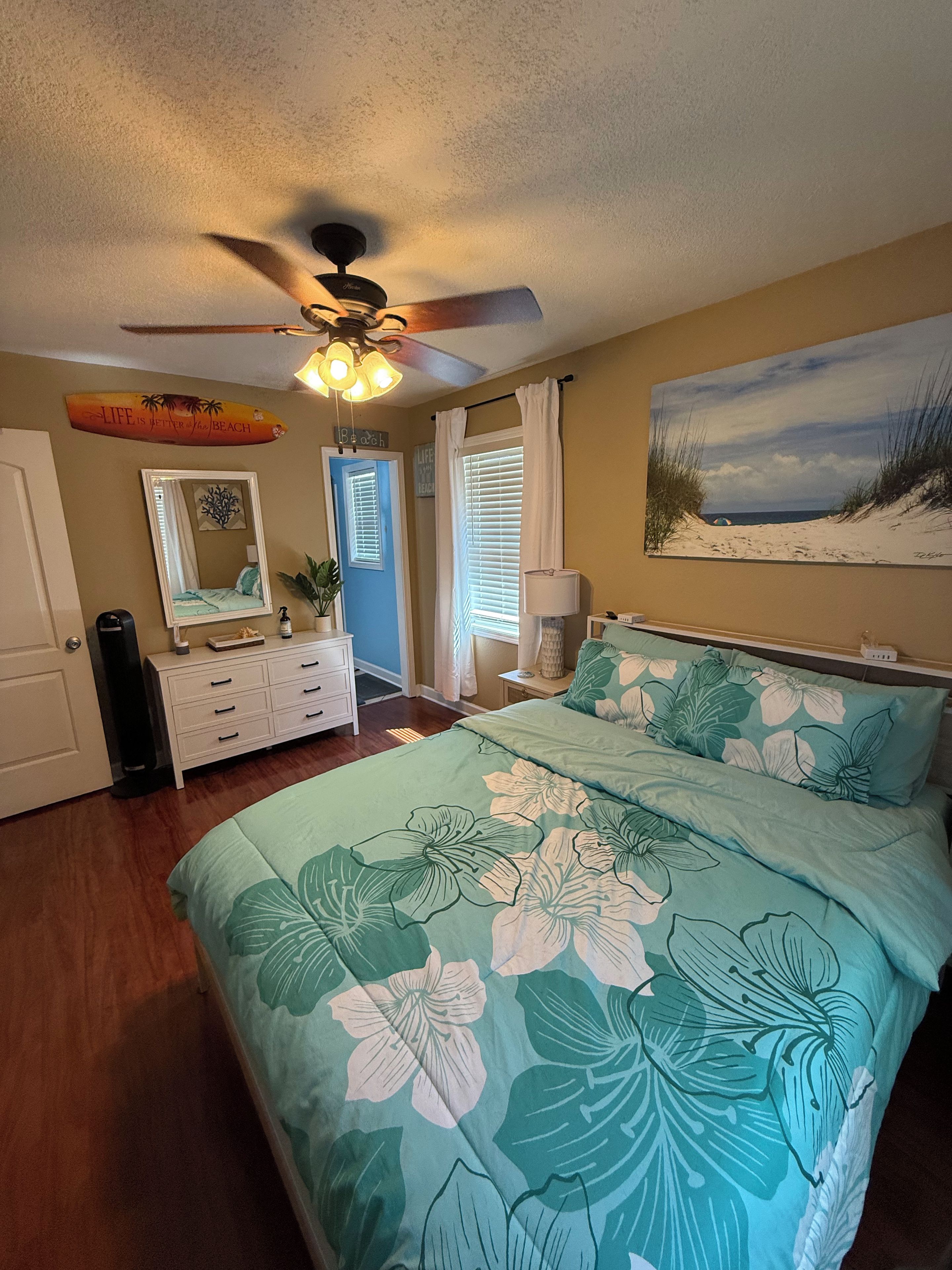 Un dormitorio con cama, ventilador de techo, puerta, cómoda, espejo, ventana con cortinas y un cuadro con motivos playeros en la pared.