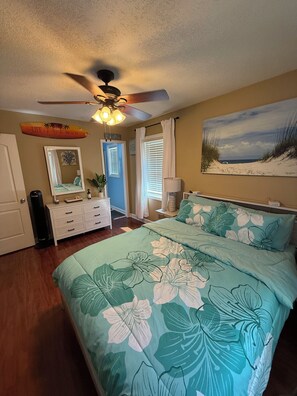 Ein Schlafzimmer mit Bett, Deckenventilator, Tür, Schminktisch, Spiegel, Fenster mit Vorhängen und einem Wandbild mit Strandmotiv.