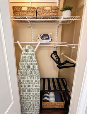 Ein schmaler begehbarer Kleiderschrank mit Regalen, einem Bügelbrett, einer Kleiderstange und einem Korb.