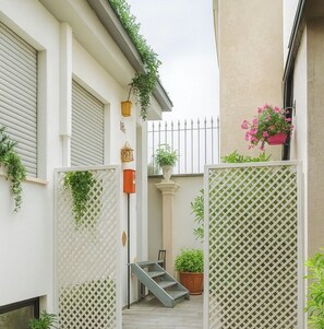 A narrow outdoor space with a white fence, potted plants, a small staircase, and a metal railing.