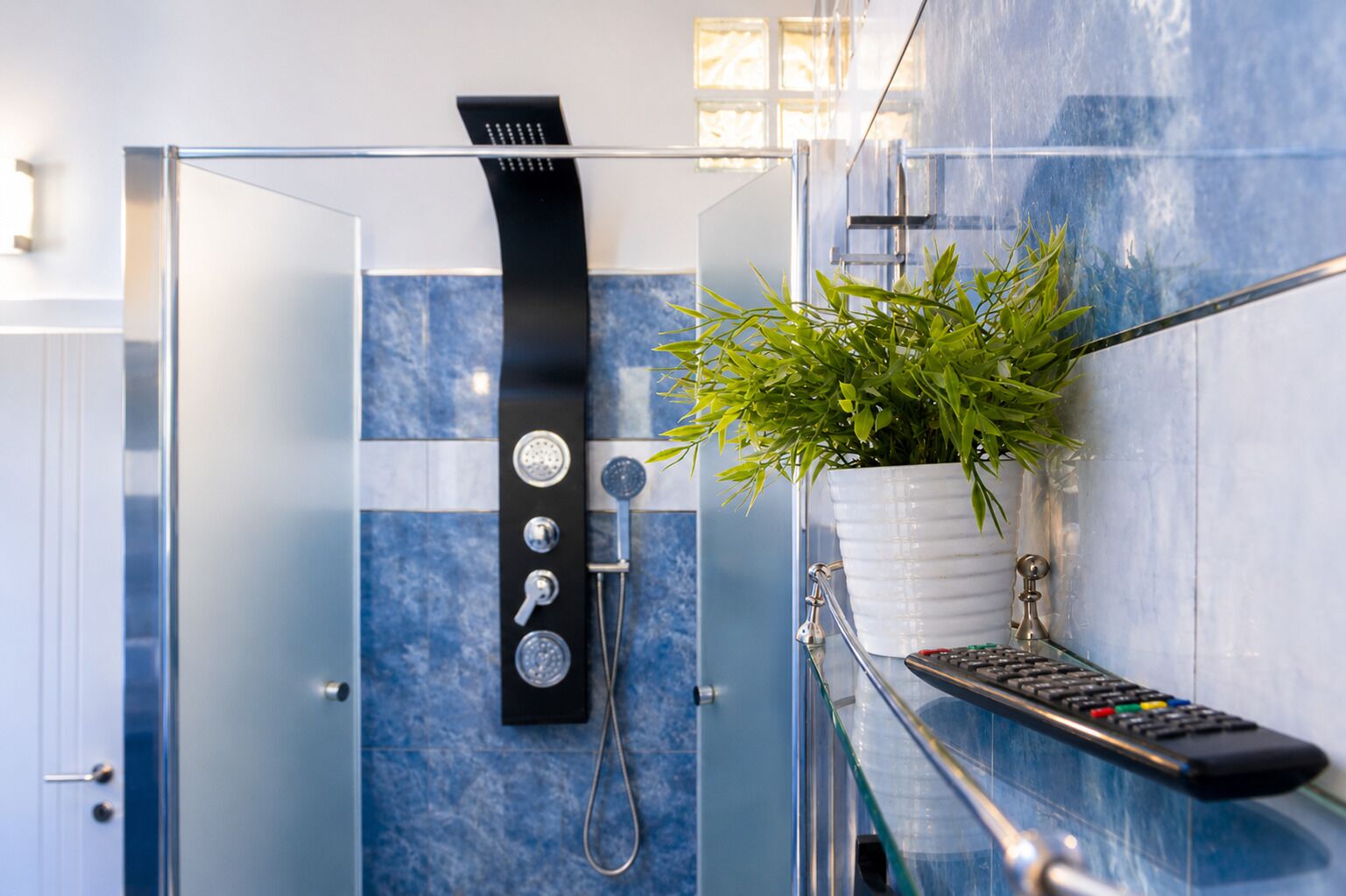 A modern bathroom with a glass-enclosed shower, a potted plant, and a remote control on a glass shelf.