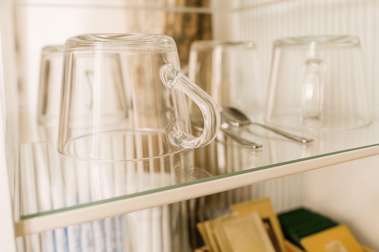 Una taza de vidrio transparente sobre una estantería con asa, un par de tijeras y una caja amarilla al fondo.