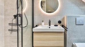 A modern bathroom with a glass shower enclosure, a round mirror with a light, a white sink with a black faucet, and a wooden vanity. - Floreal : Villa 1900 au Coeur de St Gilles (Saint-Gilles-Croix-de-Vie)