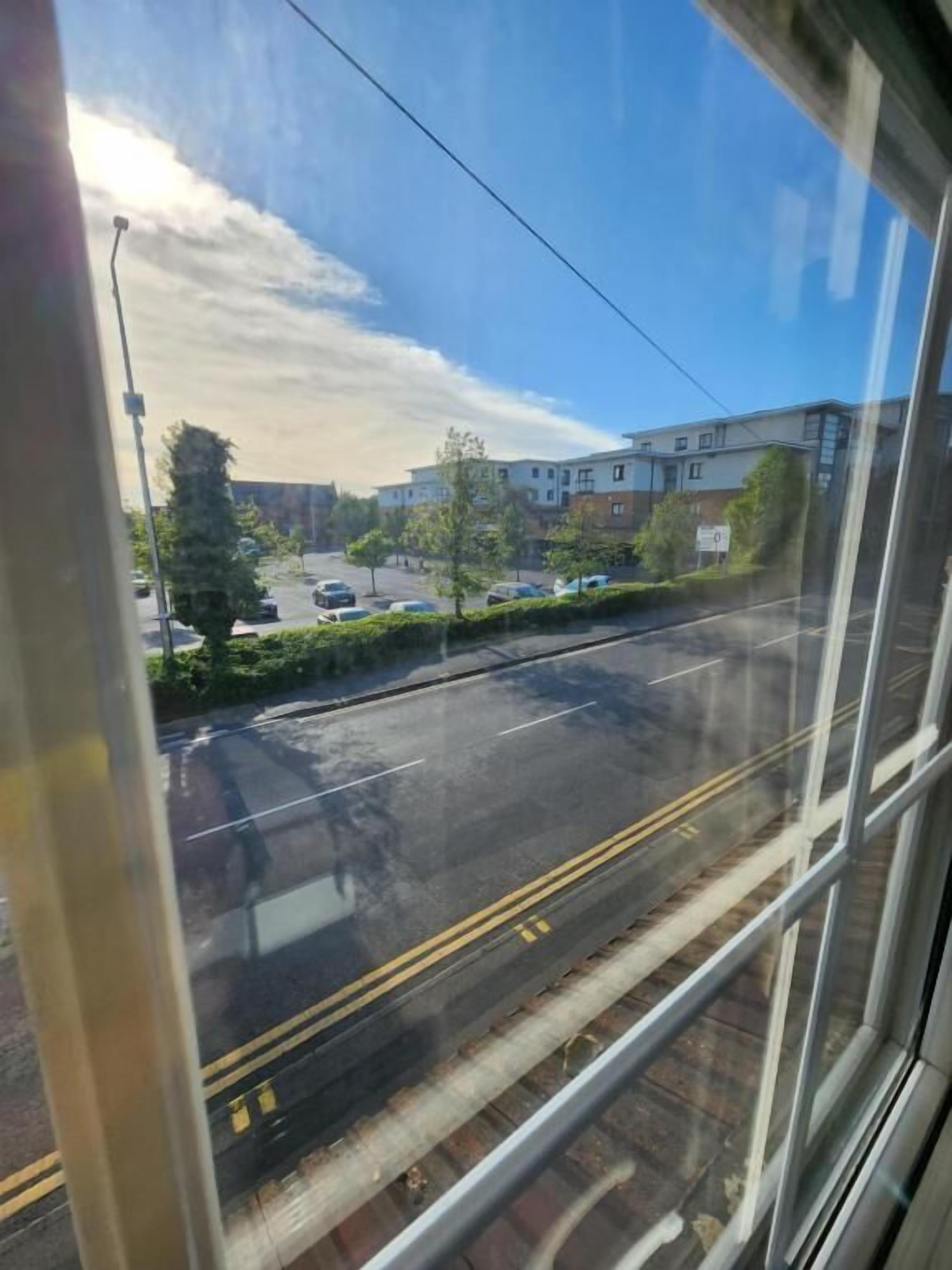 Vista a través de una ventana que muestra una calle con edificios, cielo despejado y un camino con vehículos.