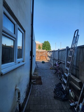 A narrow outdoor corridor with a wooden fence, a bicycle, and a ladder leaning against the wall. - The Ivy (Grays)