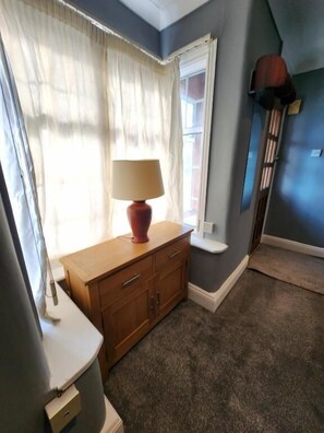 A room with a wooden cabinet, a lamp, a window with curtains, and a blue wall. - The Ivy (Grays)