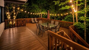 An outdoor deck with a glass-top table and chairs, surrounded by a wooden railing and string lights.