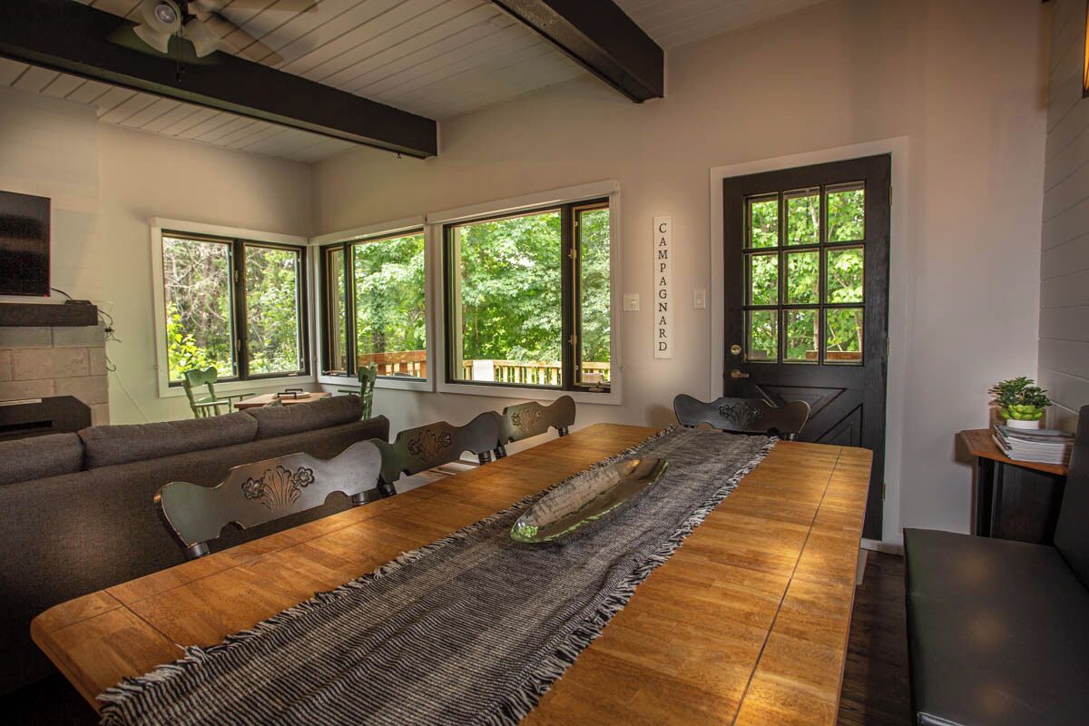 A dining area with a long wooden table, cushioned seating, and large windows offering a view of greenery.