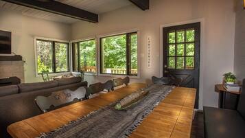 A dining area with a long wooden table, cushioned seating, and large windows offering a view of greenery.