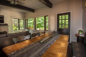 A dining area with a long wooden table, cushioned seating, and large windows offering a view of greenery.