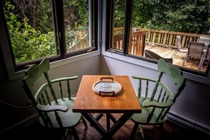 A corner room with two green chairs and a wooden table, a window with a view of a deck and greenery.