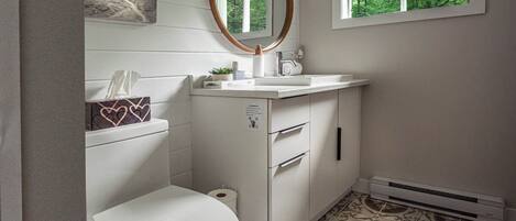 Une salle de bain comprenant des toilettes, un lavabo, un miroir et une fenêtre donnant sur des arbres.