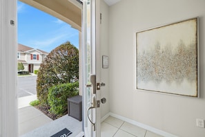A residential entrance with a door, a framed artwork on the wall, and a view of a street and houses. - Private Pool •Free Parking •Cozy Solara Resort Home Near Disney •Sleeps 12 Guest (Kissimmee)