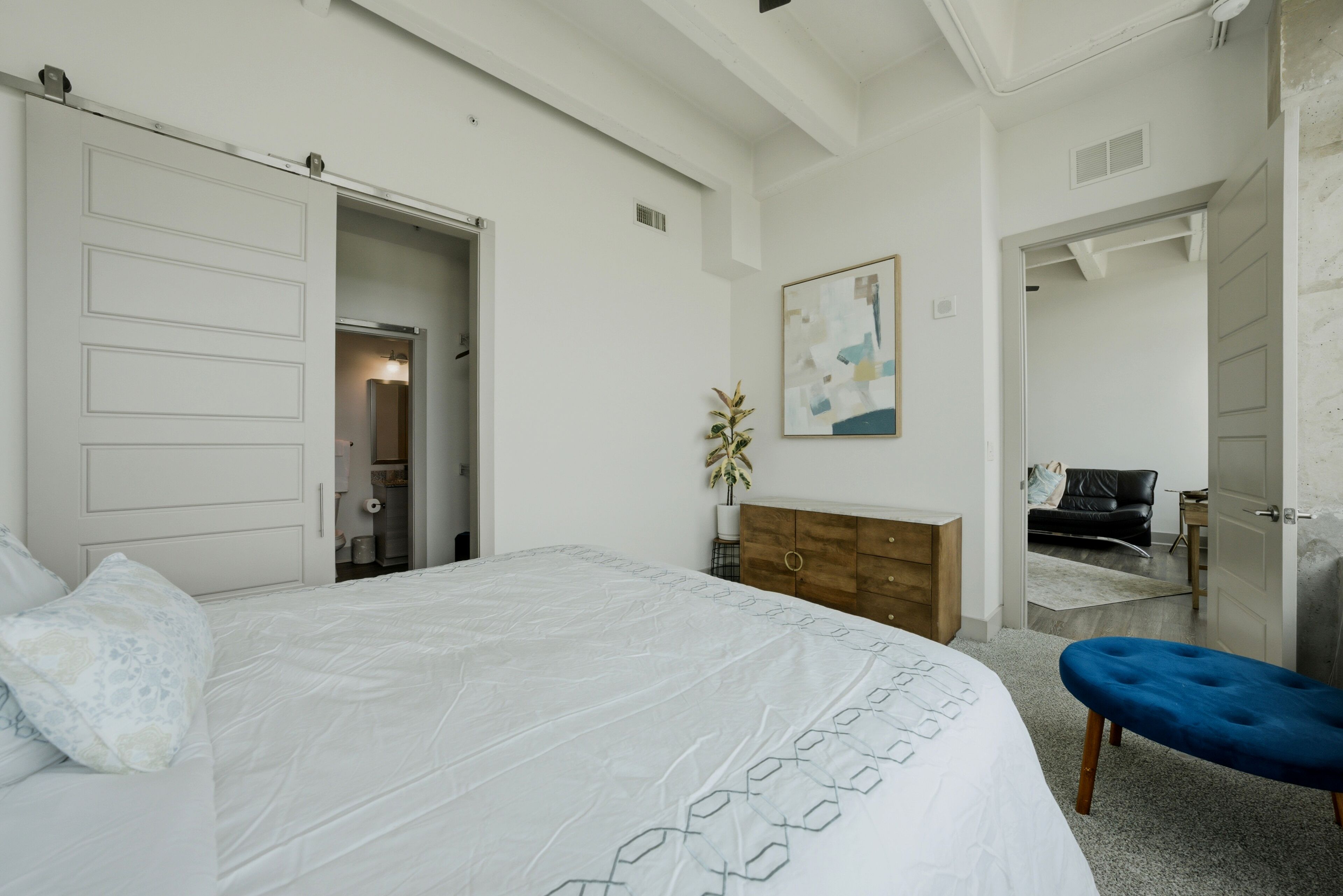 Une chambre à coucher avec un lit, un pouf bleu, une commode en bois, un tableau et une porte donnant sur une autre pièce.
