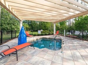 Een buitenzwembad met een pergola erboven, omringd door een verhard terras en een metalen hek.