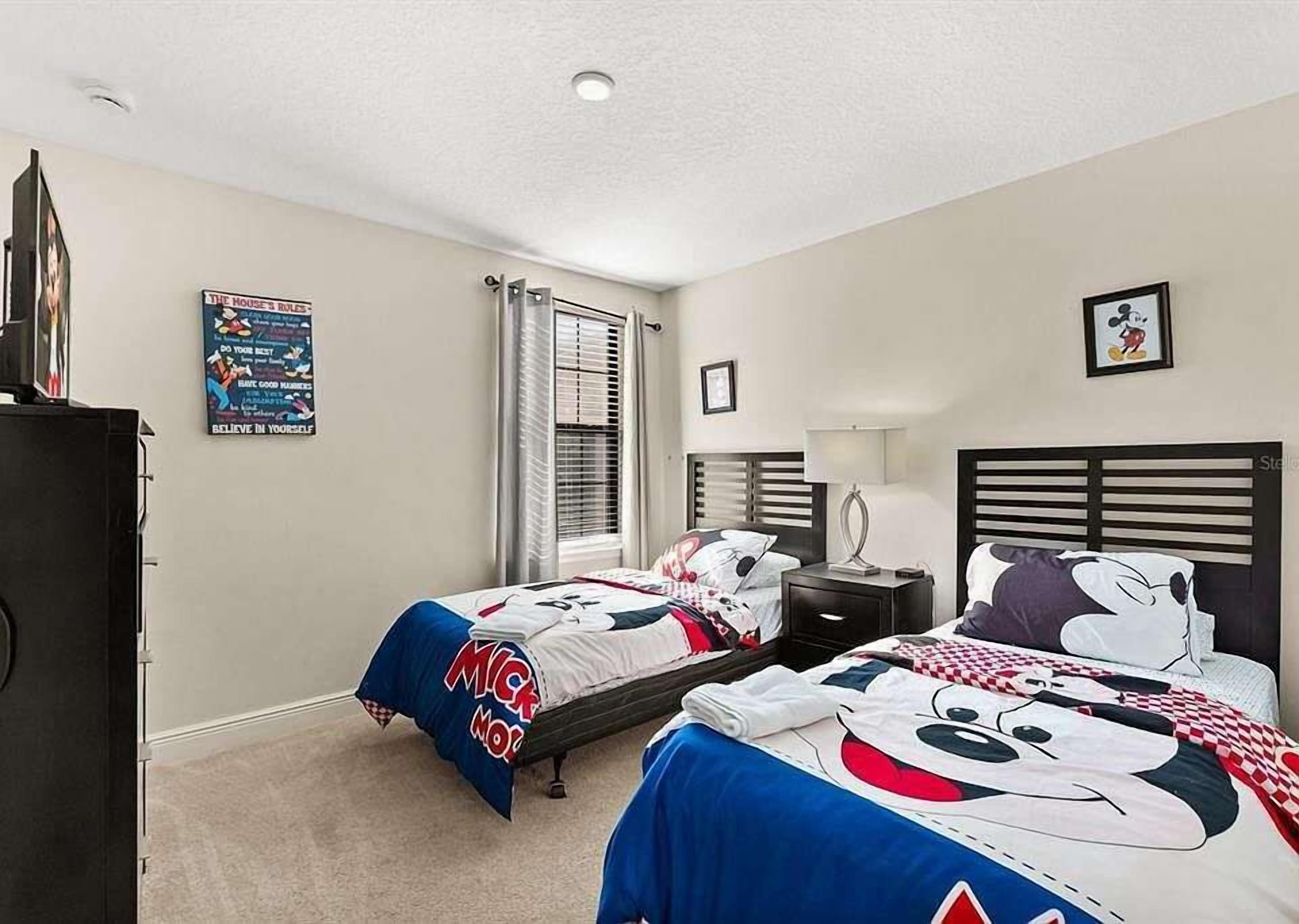 Une chambre avec deux lits, chacun agrémenté de linge de lit aux motifs de Mickey Mouse, une table de chevet, une lampe et une affiche au mur.