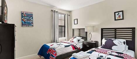 Une chambre avec deux lits, chacun agrémenté de linge de lit aux motifs de Mickey Mouse, une table de chevet, une lampe et une affiche au mur.