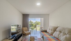 A modern living room with a flat-screen TV, a sofa, a chair, and a view of a garden through a glass door. - Beautiful apt Steps From Muta Beach Code Mut0047 (Porto Seguro)
