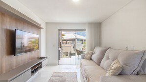 A modern living room with a flat-screen TV mounted on a wooden panel wall, a beige sofa with cushions, and a glass door leading to an outdoor area with a view of a building and greenery. - Apartment With Barbecue. in Cond. w/ Pool Mut0056 (Porto Seguro)