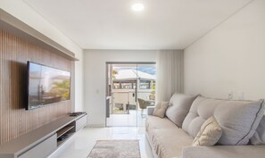 A modern living room with a flat-screen TV mounted on a wooden panel wall, a beige sofa with cushions, and a glass door leading to an outdoor area with a view of a building and greenery. - Apartment With Barbecue. in Cond. w/ Pool Mut0056 (Porto Seguro)