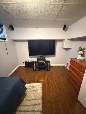 Une pièce avec un téléviseur à écran plat fixé au mur, une commode en bois, un lit recouvert d’une courtepointe bleue et un petit tapis sur le sol.