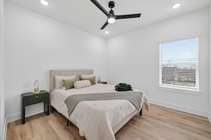 A bedroom with a bed, a nightstand, a ceiling fan, a window with blinds, and a view of the outside.
