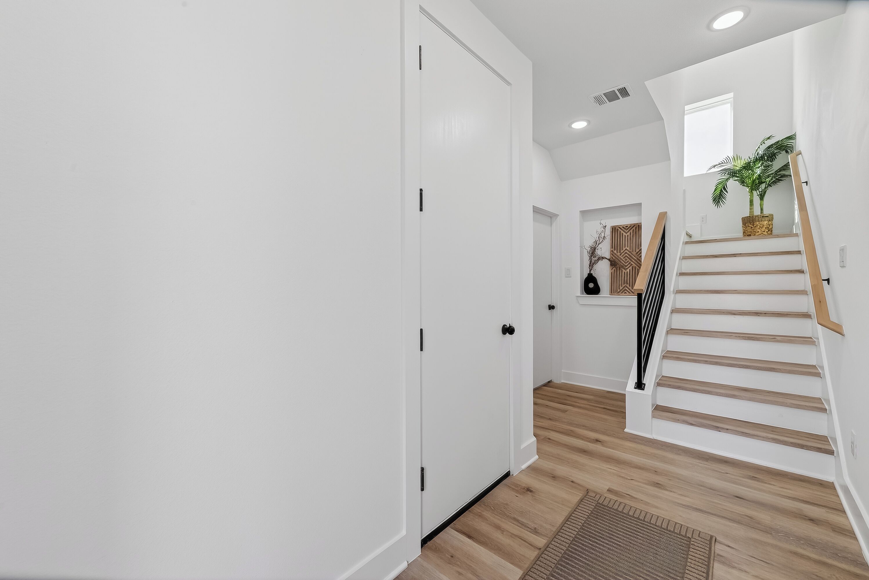A hallway with a white door, a wooden staircase, and a plant on the landing.