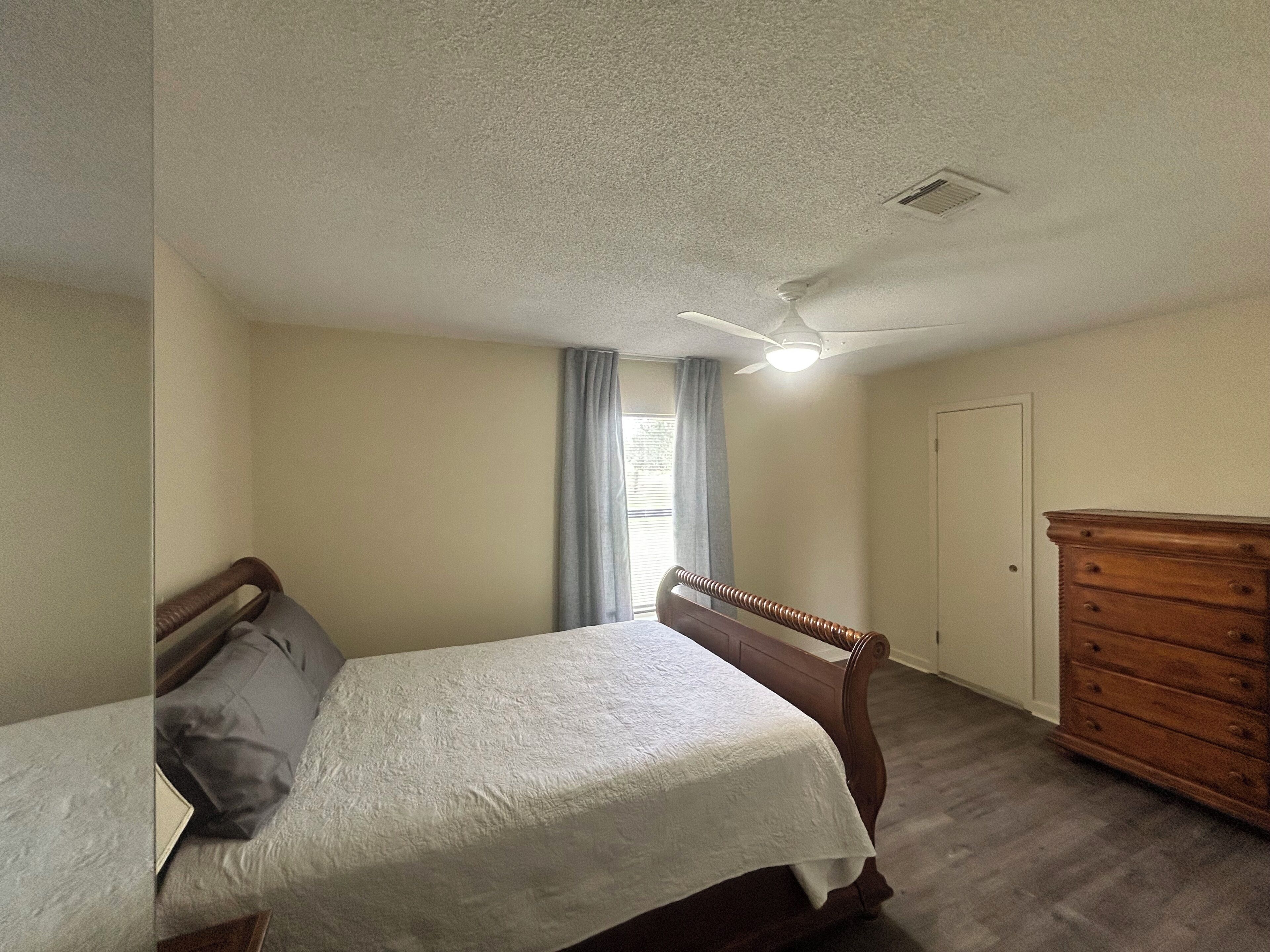 A bedroom with a bed, a dresser, a ceiling fan, and a window with curtains.