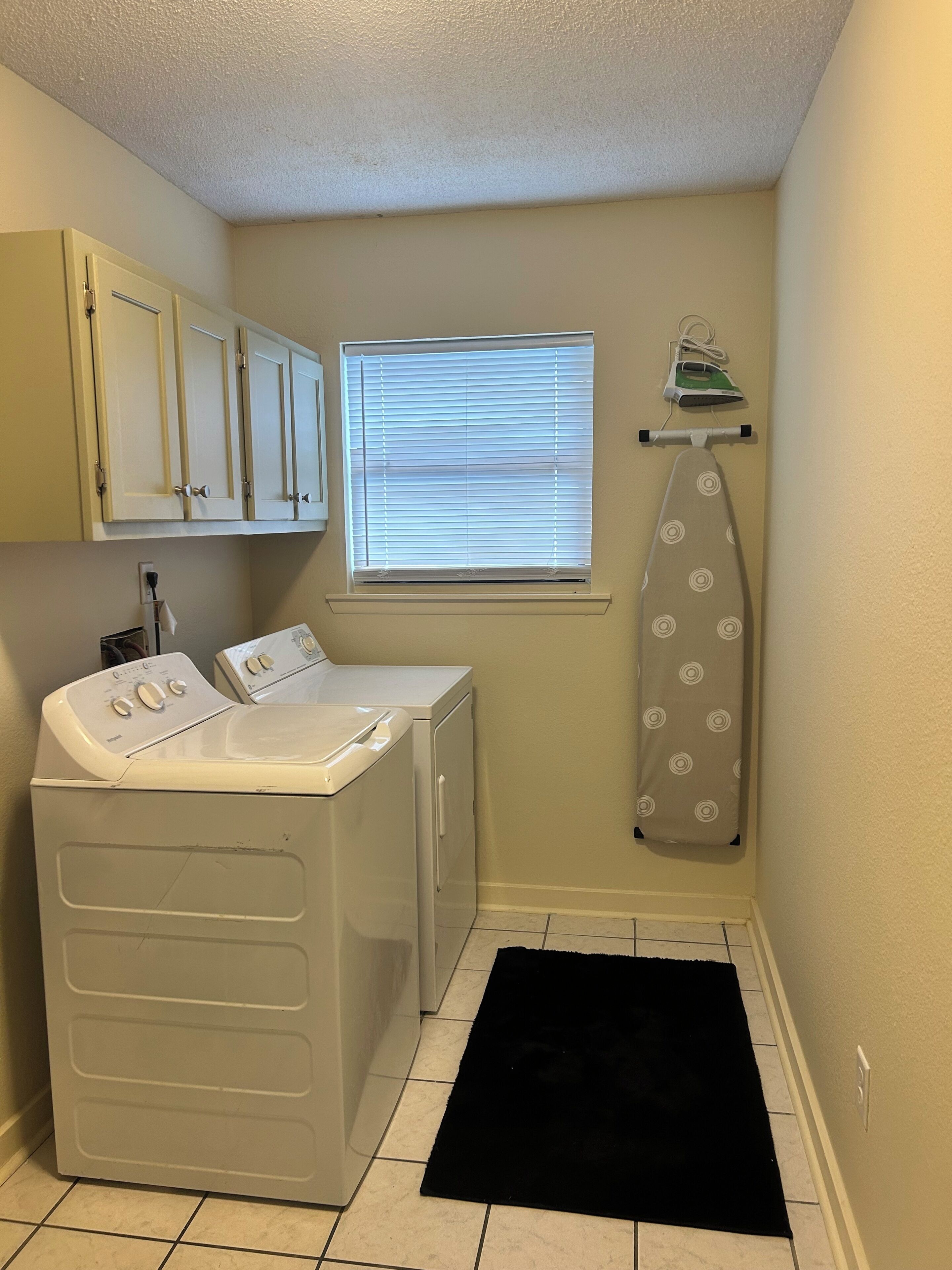 A laundry room with a washing machine, dryer, ironing board, and window with blinds.