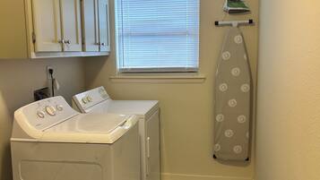 A laundry room with a washing machine, dryer, ironing board, and window with blinds.